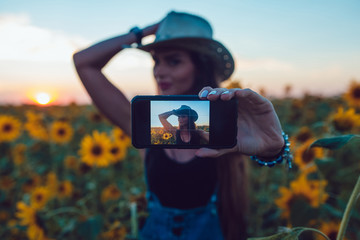 Fototapeta premium Girl in cowboy hat sitting on a suitcase in a sunflower field. Sunset.