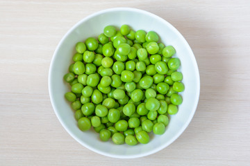 Fresh green peas in white ceramic bowl on wooden background. Top view.