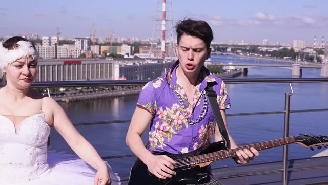 Man in skirt play electric guitar on seafront in sunny day dance cancan with girl in ballerina suit. Crazy people. Surrealism
