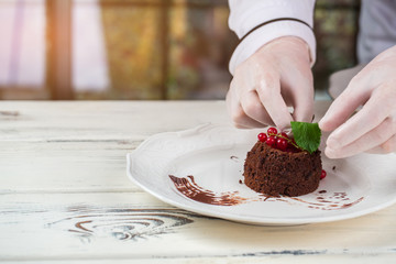 Hands touch dessert on plate. Mint and red currant. Chef prepared chocolate sponge cake. Wooden table with sweet dish.