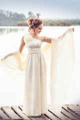 Tender bride in a white dress standing on the pier spread his hands on the background of the river.