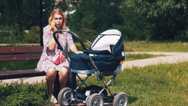 Mother sway baby carriage and speaking on phone in summer park. Sunny day. Mothercare. Motherhood