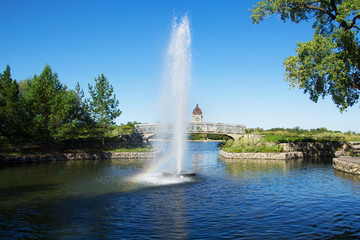 Early morning photo of a fountain in Regina Saskatchewan's Wascana Park with a rainbow in the spray. Landscape view with a footbridge and the provincial legislature in the distance. - obrazy, fototapety, plakaty