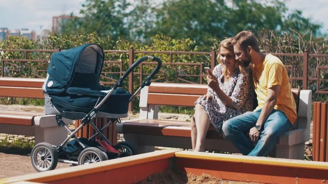 Young parents with baby carriage on bench. Using smartphone. Family. Playground. Summer sunny day