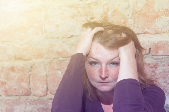 Stressed redhead young woman is holding her head by both hands and is looking at the camera. Old brick wall is in the background.