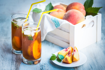 Glasses with homemade ice tea, peach flavored. Freshly cut peach slices for arrangement. White crate full with peaches in the background.