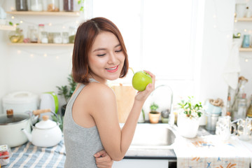 Obraz na płótnie Canvas Happy Young Asian Woman Eating Green Apple on Kitchen. Diet. Die