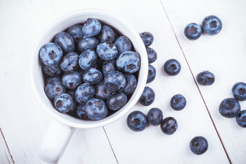 View from above on handful of ripe and fresh blueberries in white cup and some blueberries sprinkled on white wooden desk
