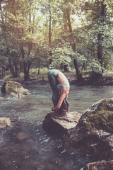 Attractive young woman doing stretching exercises in beautiful untouched nature environment. She standing on the huge rock in the middle of cold mountain river.