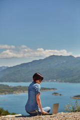 Fototapeta premium Beautiful young freelancer woman using laptop sitting on the lake.Happy smiling girl working online.Studying and learning using notebook computer.Freelance work,business people concept.