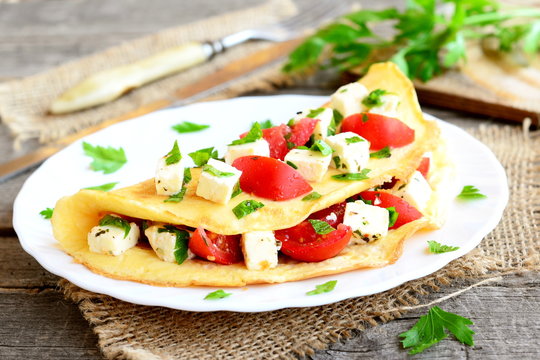 Homemade stuffed omelet on a plate. Egg omelet stuffed with fresh tomatoes, cheese and green parsley. Healthy vegetarian breakfast recipe. Fork, knife, cutting board on wooden background. Closeup