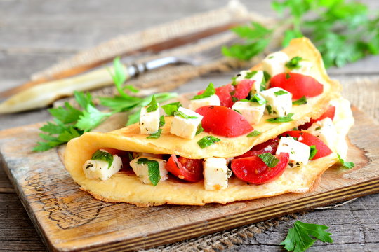 Delicious stuffed omelette on a wooden board. Fried egg omelette with a filling of cherry tomatoes, cheese and parsley. Easy cooking breakfast. Fork, knife, parsley sprigs on wood background. Closeup