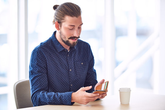 Trendy Handsome Man With Long Hair Tied Back In A Bun Busy Using His Mobile Phone To Stay Connected While Sitting At Restaurant Table.