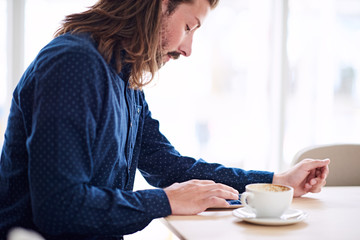 landscape image of handsome caucasian man with long brown hair using tablet that is lying on the table in front of him next to his coffee.
