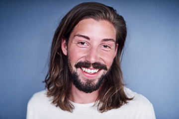 Young white man with long loose brown hair and a beard standing in front of a blue background while wearing a white t-shirt with a positive aura.