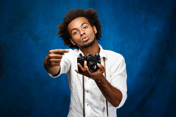 Young handsome african man holding old camera over blue background.