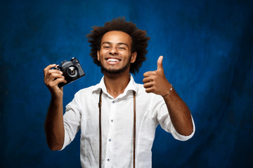 Young handsome african man holding old camera over blue background.