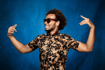 African man listening music in headphones, dancing over blue background.