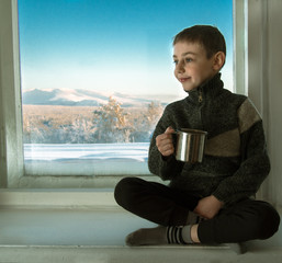 Toned image of a little boy sitting on an old window sill next to the window and holding in his hand a metal cup against the background of the winter mountains and blue sky