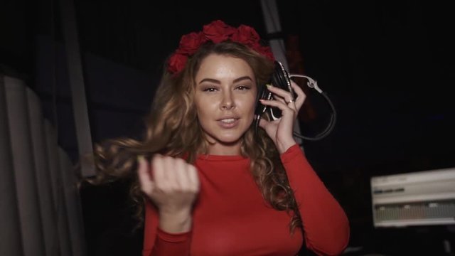 Dj girl in red dress with rim on head touch face by hand at turntable in nightclub. Smile in camera. Headphones. Slow motion