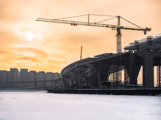 Toned image under construction Road Bridge over the frozen river with a large tower crane against the backdrop of a cloudy sky with the sun and residential buildings