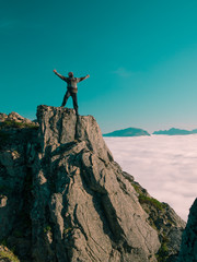 Toned image adult man with backpack stands on the edge of a cliff and screams and shows his hand into the distance against the blue sky and thick clouds floating down