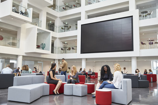 Students sit talking under AV screen in atrium at university