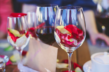Beautiful glass with rose leafs, rose-petal, rose petals on a romantic date with wine, candles in candlelight and with celebrating couple on the background, interior for a romantic date diner 