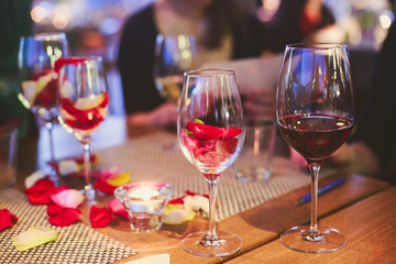 Beautiful glass with rose leafs, rose-petal, rose petals on a romantic date with wine, candles in candlelight and with celebrating couple on the background, interior for a romantic date diner 