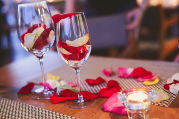 Beautiful glass with rose leafs, rose-petal, rose petals on a romantic date with wine, candles in candlelight and with celebrating couple on the background, interior for a romantic date diner 