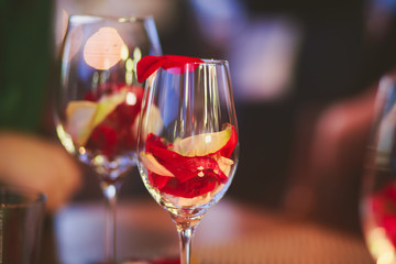 Beautiful glass with rose leafs, rose-petal, rose petals on a romantic date with wine, candles in candlelight and with celebrating couple on the background, interior for a romantic date diner 