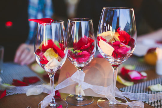 Beautiful glass with rose leafs, rose-petal, rose petals on a romantic date with wine, candles in candlelight and with celebrating couple on the background, interior for a romantic date diner 