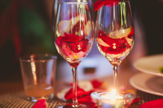 Beautiful glass with rose leafs, rose-petal, rose petals on a romantic date with wine, candles in candlelight and with celebrating couple on the background, interior for a romantic date diner 