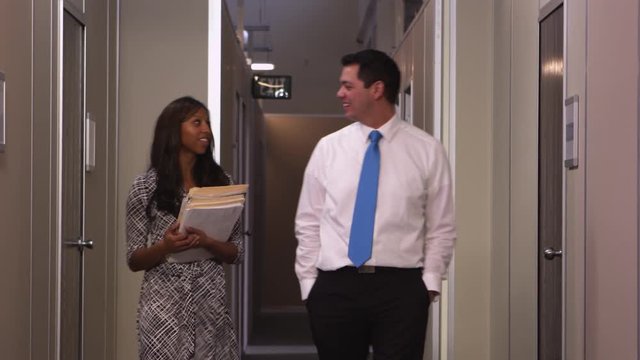 A business man and woman walk down the hallway towards the camera as they talk about work