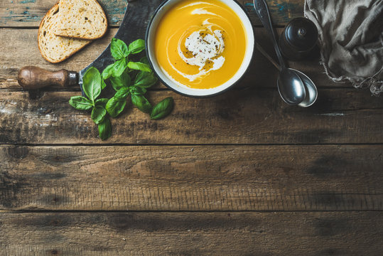 Pumpkin cream soup in bowl with fresh basil, spices and grilled bread slices over old rustic wooden background, top view, copy space, horizontal composition