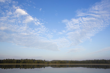 A beautiful morning by the lake. Forest is reflecting from the water. Clouds are like heart shaped in the blue sky.