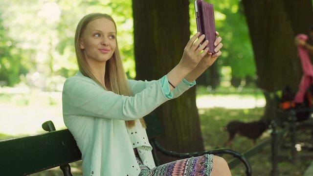 Pretty girl doing selfies on tablet while sitting in the park
