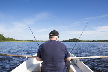 An elderly man is fishing on a rowing boat. He is using two fishing rods while rowing the boat.