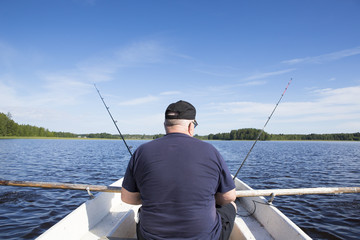An elderly man is fishing on a rowing boat. He is using two fishing rods while rowing the boat.