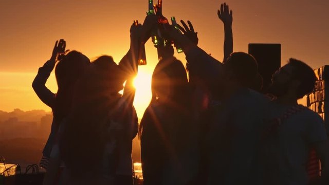 Silhouettes of young people toasting with beer bottles and dancing with raised arms to the music played by dj at rooftop party during beautiful city sunset