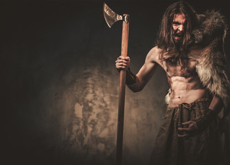 Obraz na płótnie Canvas Angry viking with ax in a traditional warrior clothes, posing on a dark background.