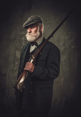 Fototapeta na wymiar Senior hunter with a shotgun in a traditional shooting clothing, posing on a dark background.