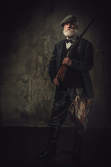Fototapeta premium Senior hunter with a shotgun in a traditional shooting clothing, posing on a dark background.