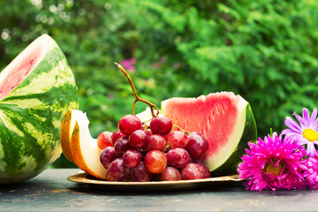 cut slices of ripe yellow melon, watermelon, a bunch of grapes and flowers asters on a table with natural green background. shallow depth of field