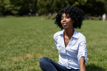 Black woman in a park sitting on lawn happy
