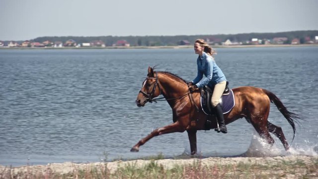 Tracking of brown colored horse with leaning ecstatic female rider on it speeding through lake water in slow motion