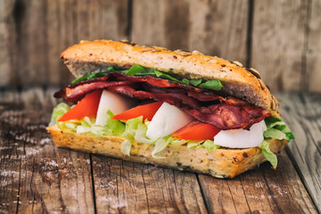 Bacon baguette with egg, tomato and lettuce. On an old wooden table.