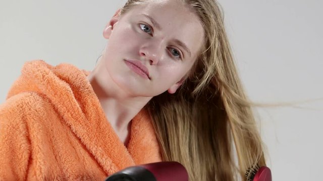 Face of blonde teenage girl wearing orange bathrobe blow drying her wet long hair with hairdryer after shower in bathroom closeup