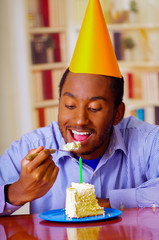 Charming man wearing blue shirt and hat sitting by table eating piece of cake in front, looking happy, celebrating alone concept