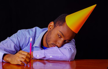 Bored looking man wearing blue shirt and hat sitting by table holding a single candle burning, sad expression facing camera, celebrating alone concept